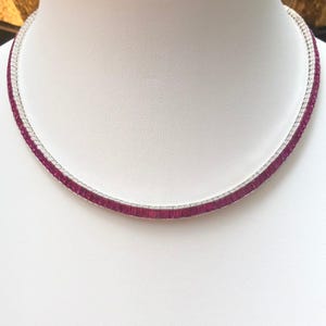 Puede incluir: Un collar plateado con una fila de piedras preciosas rectangulares de color rosa intenso. Las piedras están engastadas muy juntas, creando una línea continua de color. El collar se exhibe sobre una superficie blanca.