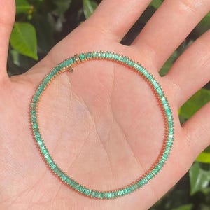 Puede incluir: Un brazalete dorado con una fila continua de piedras preciosas rectangulares de color verde esmeralda. El brazalete se sostiene en una mano, con un fondo verde borroso.