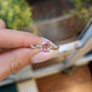 Puede incluir: Un anillo de oro con una gran gema rosa ovalada. La banda tiene un diseño retorcido con pequeñas piedras claras. El anillo se sostiene contra un fondo borroso, destacando los detalles. El diseño del anillo es intrincado.