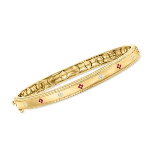 Puede incluir: Un brazalete dorado con cierre de bisagra. El brazalete está decorado con pequeñas piedras preciosas rojas y transparentes en un patrón floral. Tiene una superficie texturizada y una serie de recortes circulares a lo largo del borde interior.