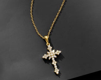 May include: A gold-colored necklace with a pendant in the shape of a cross. The cross is adorned with multiple clear, sparkling gemstones. The chain is delicate and hangs against a dark background, highlighting the pendant's intricate design.
