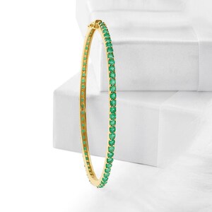 May include: A gold-colored bangle bracelet with a row of vibrant green gemstones. The bracelet is set against a white background, highlighting the contrast between the gold and green. The gemstones are evenly spaced around the bracelet.