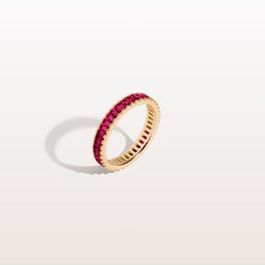 May include: A gold ring featuring a continuous line of rectangular, bright red gemstones. The ring is set against a white backdrop, with a soft shadow. This jewellery item is a classic and elegant design.