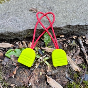 May include: Two neon green zipper pulls with red cords are shown against a natural background of leaves and a rock. The pulls have a rounded rectangular shape with horizontal lines. The red cords are looped together at the top.