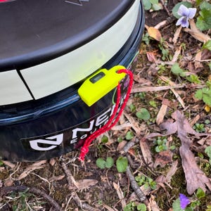 May include: A close-up of a yellow safety whistle attached to a red cord. The whistle is attached to a black and white container with the word "NE" in white letters.