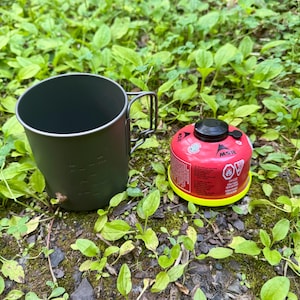 Op de afbeelding: Een donkergrijze kampeermok met een opvouwbaar handvat staat naast een rood-gele MSR brandstofcartridge. De mok heeft maatstreepjes. De items staan op groene planten en aarde, wat duidt op gebruik buitenshuis.