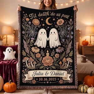 Puede incluir: Una manta tejida negra con dos fantasmas blancos tomados de la mano, rodeados de motivos florales, setas, telarañas y una luna creciente. El texto dice 'Til death do us part', 'Julia & Daniel' y '16.10.2025' en una cinta.