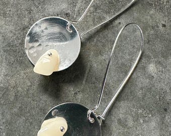 Puede incluir: Un par de pendientes de plata con un colgante de piedra blanca. Los pendientes están hechos de un disco de metal martillado con una pequeña perla de plata en la piedra.