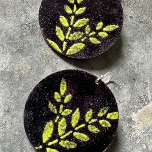 May include: A pair of handmade earrings. Each earring is a dark purple, circular disc with a textured surface. A bright yellow-green leaf design is painted on each disc. The earrings have silver-colored ear wires.
