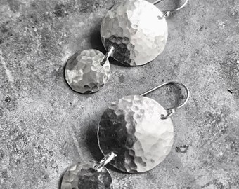 Puede incluir: Un par de pendientes de plata con textura martillada. Cada pendiente tiene dos discos redondos, uno más grande que el otro.