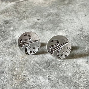 May include: Two silver cufflinks with a design of a needle and thread on a spool of thread.