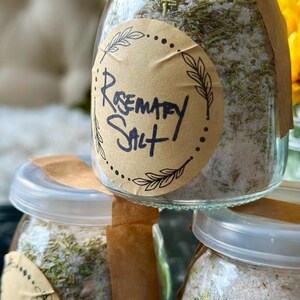 Puede incluir: Tres frascos de vidrio llenos de una mezcla de sal y hierbas, etiquetados como "Rosemary Salt". Los frascos tienen tapas de papel marr&oacute;n y est&aacute;n apilados uno encima del otro.