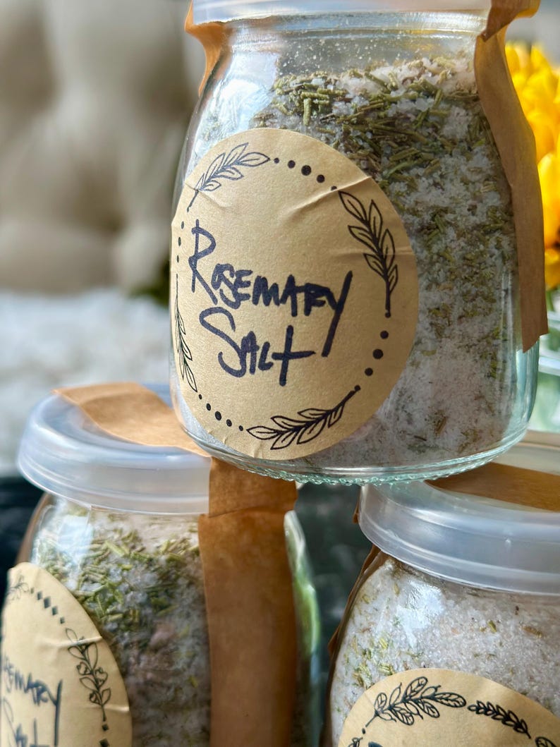 Puede incluir: Tres frascos de vidrio llenos de una mezcla de sal y hierbas, etiquetados como "Rosemary Salt". Los frascos tienen tapas de papel marr&oacute;n y est&aacute;n apilados uno encima del otro.