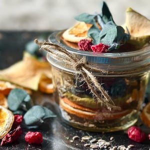 Może przedstawiać: Szklany słoik wypełniony suszonymi owocami, ziołami i kwiatami. Słoik jest związany sznurkiem i ma metalową pokrywkę. Słoik stoi na ciemnej powierzchni, wokół której rozrzucone są inne suszone owoce i zioła.