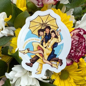 以下が含まれることがあります： 黄色い傘の下で踊る3人の漫画のイラストが描かれたステッカーで、「Singing in the Rain」の文字が入っています。ステッカーは白い縁取りで、色とりどりの花を背景にしています。