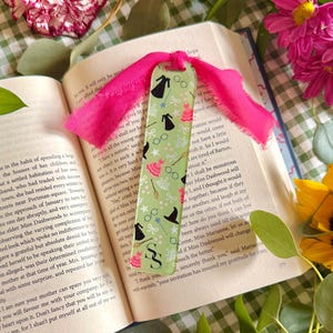 May include: A green bookmark with a pink ribbon, featuring a pattern of black witch hats, dresses, and other magical symbols. The bookmark is placed in an open book with text.