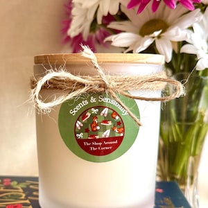 May include: A frosted glass candle jar with a wooden lid, tied with a jute twine bow. A circular green label features the text Scents & Sensibility and The Shop Around The Corner, with illustrations of foxes, books, and ballet slippers.