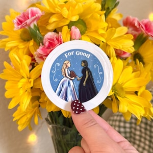 May include: A round sticker with the text "For Good" and illustrations of two women in gowns holding hands. The sticker is held in front of a bouquet of yellow and pink flowers.