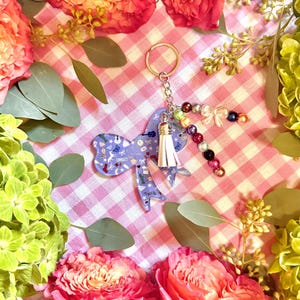 Puede incluir: Un llavero morado en forma de lazo con una borla blanca y un adorno de cuentas. El lazo presenta un patr&oacute;n de instrumentos musicales y otros peque&ntilde;os dise&ntilde;os. El llavero est&aacute; rodeado de flores rosas y rojas y follaje verde.