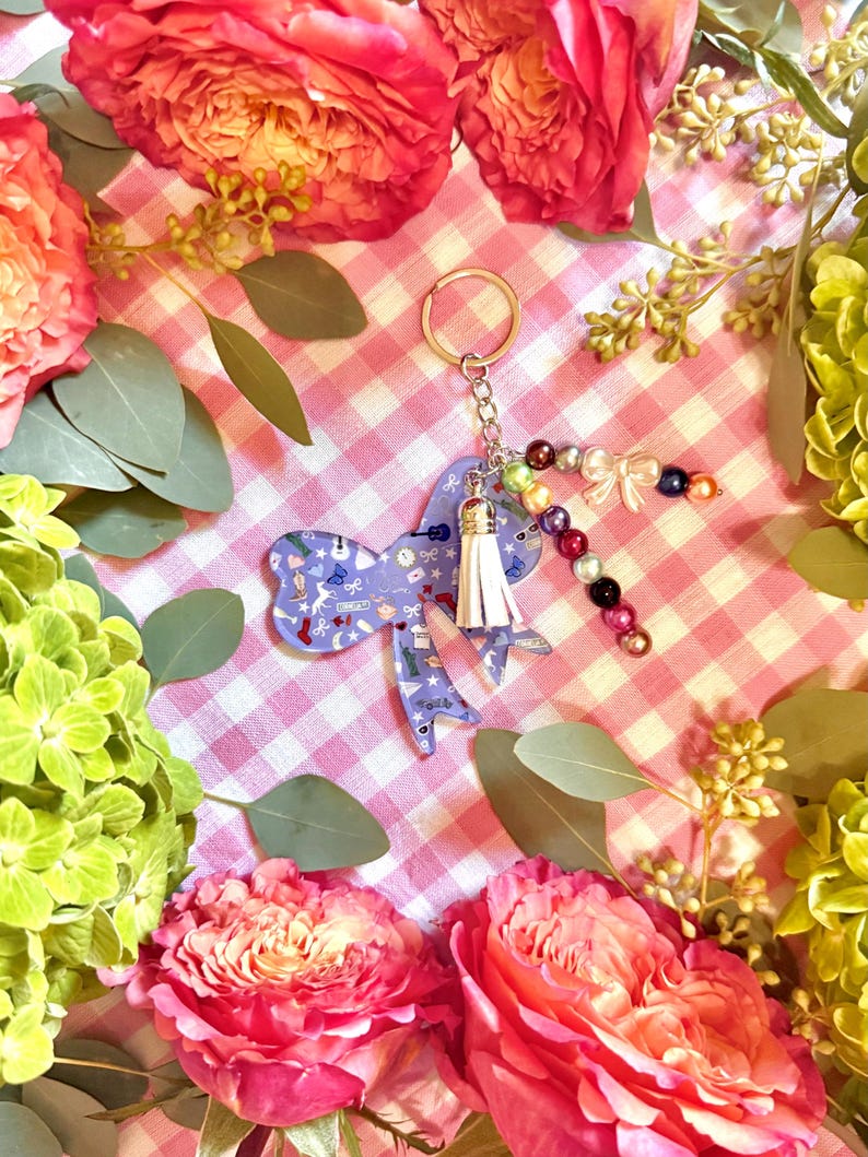 Puede incluir: Un llavero morado en forma de lazo con una borla blanca y un adorno de cuentas. El lazo presenta un patr&oacute;n de instrumentos musicales y otros peque&ntilde;os dise&ntilde;os. El llavero est&aacute; rodeado de flores rosas y rojas y follaje verde.