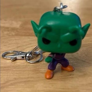 May include: A green Piccolo keychain figure with black eyes, orange shoes, and purple pants. The figure has a silver keychain clasp attached. The figure is standing on a wooden surface.