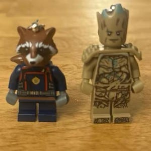 May include: Two Lego minifigures: Rocket Raccoon in a blue and red outfit and Groot in a tan and brown outfit. Rocket has a raccoon head and Groot has a tree-like appearance. Both figures are standing on a wooden surface.