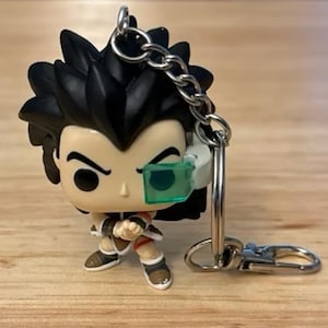May include: A small, stylized figurine keychain of a character with black spiky hair, a white and green eye visor, and a determined expression. The figure is wearing a brown and white outfit. The keychain has a silver chain and clasp.