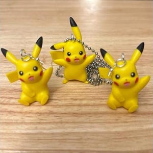 Może przedstawiać: Trzy żółte zawieszki Pikachu, jedna na srebrnym łańcuszku i dwie jako kolczyki. Zawieszki są wykonane z plastiku i mają czarne oczy i czerwone policzki.
