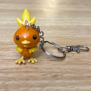 May include: Orange and yellow Torchic keychain. The Torchic figure has black eyes, a yellow flame-like crest, and yellow feathers around its neck. The keychain includes a silver-colored split ring and a lobster clasp.