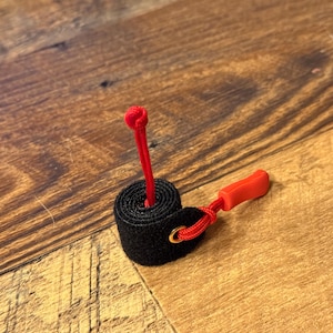 May include: A black cable tie with a red cord and a red plastic clip. The cable tie is wrapped around itself and the cord is threaded through the tie.