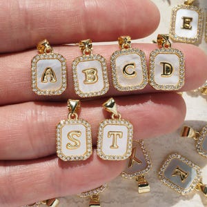 May include: Gold-toned initial pendants with a white background and a border of small, clear crystals. The letters A, B, C, D, S, and T are visible. The pendants are rectangular with a gold bail.