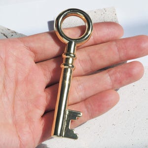 May include: A gold-colored key with a circular top and a detailed shaft. The key is held in a hand, showcasing its polished finish and intricate design. The key's teeth are clearly visible, highlighting its functionality.