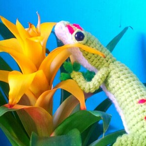 May include: A green crocheted gecko with pink spots is perched on a yellow flower with green leaves. The gecko has a white belly and a pink and white face.