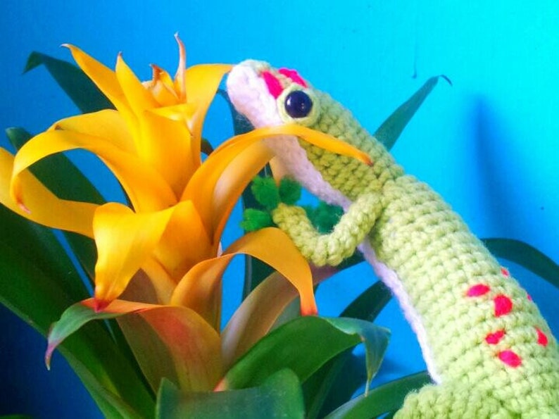 May include: A green crocheted gecko with pink spots is perched on a yellow flower with green leaves. The gecko has a white belly and a pink and white face.