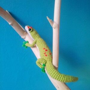 May include: Green crocheted gecko with red spots, perched on a branch against a blue background.