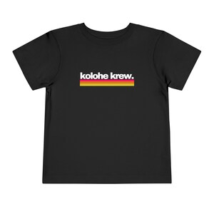 May include: Black toddler t-shirt with the text "kolohe krew" in white, above a horizontal stripe of red, yellow, and white. The shirt is made of a soft material.