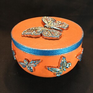 May include: Orange ceramic trinket box with a blue trim and a gold butterfly design. The box is decorated with smaller butterflies around the sides.
