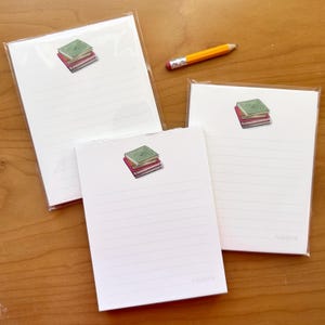 May include: Three white notepads with horizontal lines and a small illustration of a stack of books in green, red, and pink. A yellow pencil with a pink eraser rests nearby. The notepads are in clear plastic packaging.