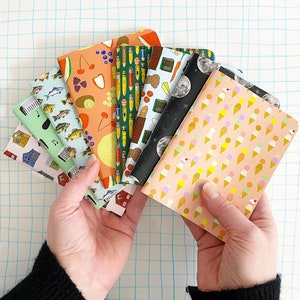 May include: A collection of eight colorful notebooks with various patterns and designs. The notebooks feature images of food, nature, and geometric shapes. The notebooks are arranged in a fan shape, with the covers facing the viewer.