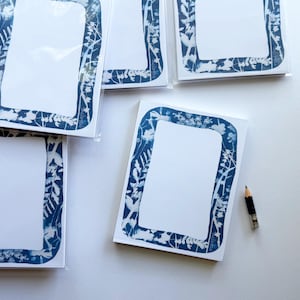 May include: A collection of rectangular notepads featuring a white center and a dark blue border decorated with a white botanical print of various leaves and stems. A small, sharpened wooden pencil with a silver ferrule lies next to the notepads.