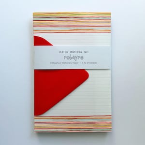 May include: A letter writing set featuring stationery paper with horizontal multicolored stripes at the top and bottom edges. A white paper band wraps around the center, printed with the text LETTER WRITING SET robayre 8 Sheets of Stationery Paper 4 A2 envelopes, partially covering a bright red envelope.