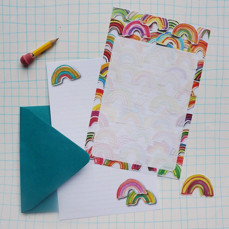 May include: A stationery set featuring rainbow-themed designs. The set includes lined stationery paper, a teal envelope, and rainbow stickers. A yellow pencil with a pink eraser is also present. The paper has a rainbow border and a rainbow pattern.