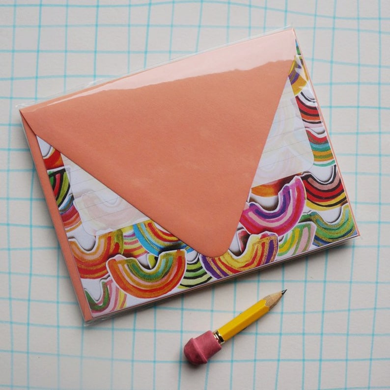 May include: A stationery set featuring a peach-colored envelope and a notepad with a colorful rainbow design. A yellow pencil with a pink eraser rests beside the stationery. The set is packaged in clear plastic.