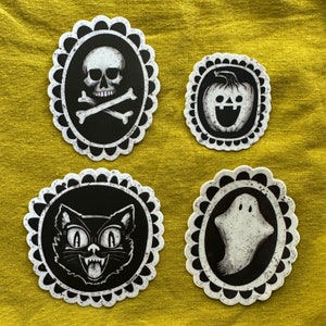 May include: Four black and white Halloween themed stickers with white scalloped borders. The stickers feature a skull and crossbones, a jack-o-lantern, a black cat, and a ghost.