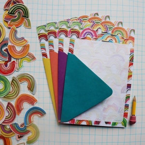 May include: A collection of colorful stationery items, including notebooks with rainbow-themed edges and covers in yellow, orange, purple, and teal. Loose rainbow stickers and a pencil with an eraser are also present.