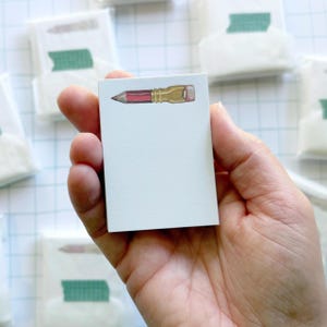 May include: A rectangular white notepad featuring a small, centered illustration of a pencil with a red body and a gold-colored metal ferrule at the top. The notepad is plain white below the pencil graphic, providing a clean space for writing notes.