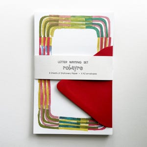 May include: A letter writing set featuring 8 sheets of stationery paper and 4 A2 envelopes. The paper has a rectangular border of painted green, pink, and yellow lines. A white paper band reads LETTER WRITING SET robayre 8 Sheets of Stationery Paper 4 A2 envelopes, with a red envelope tucked underneath.