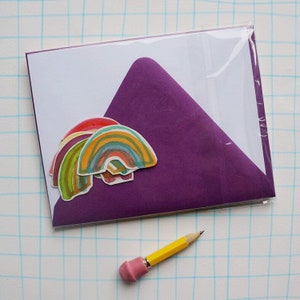 May include: A stationery set featuring a white card and a purple envelope, sealed in clear plastic. The card is decorated with colorful rainbow stickers. A yellow pencil with a pink eraser is in the foreground.