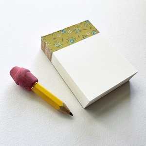 May include: A yellow pencil with a pink eraser rests beside a small notepad. The notepad has a stack of white pages and a decorative top with a green floral pattern. The pencil is sharpened and ready for use.
