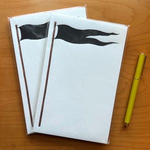 May include: Two white notepads with a black flag design, packaged in clear plastic. A yellow pen rests on a wooden surface next to the notepads. The flag illustration features a black flag on a brown pole.